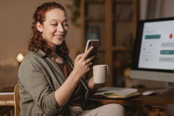 mulher-em-casa-usando-smartphone-na-frente-computador-enquanto-toma-cafe